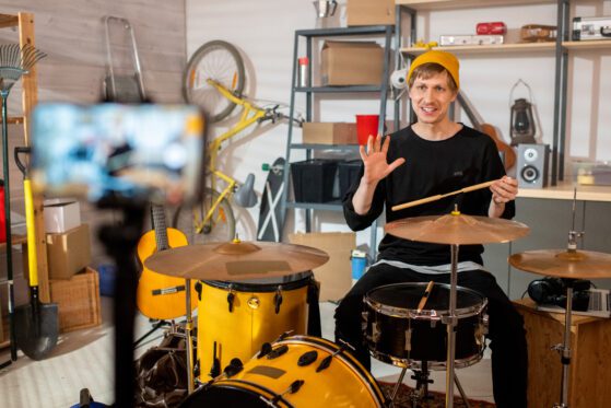 Happy young man with drumsticks sitting by drumset in front of smartphone camera for online drum classes