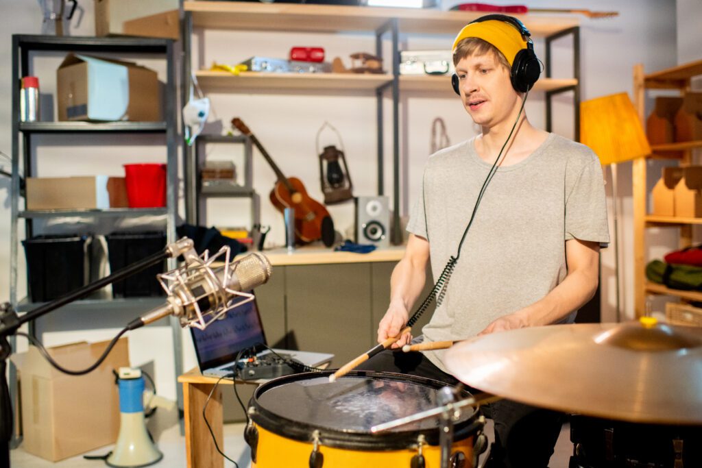 Young drummer, in his online drum class with headphones beating drum with drumsticks during individual rehearsal and recording his music in front of microphone