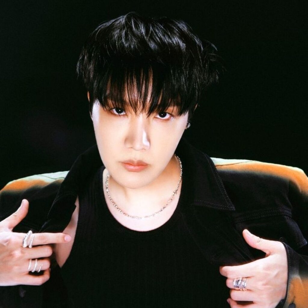 A member of a K-pop band with dark hair and intense expression, wearing a black outfit with silver rings and necklace, posing against a dark background.
