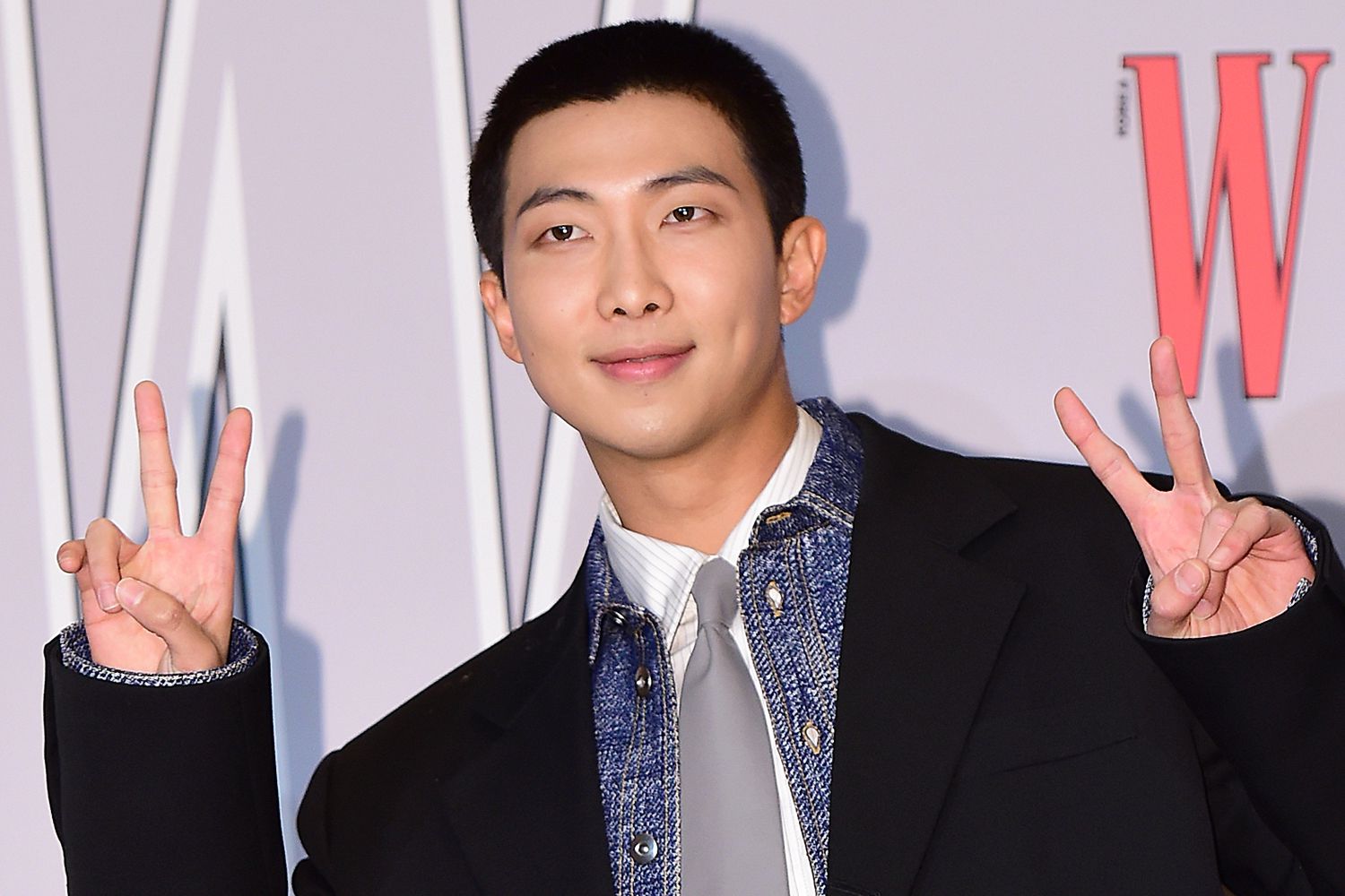 A member of a popular K-pop band posing at a W Korea event, wearing a stylish outfit and showing a peace sign with both hands.