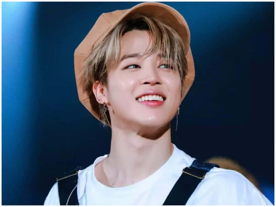 A smiling member of a K-pop band wearing a beige beret, white t-shirt, and suspenders, looking cheerful under stage lights.
