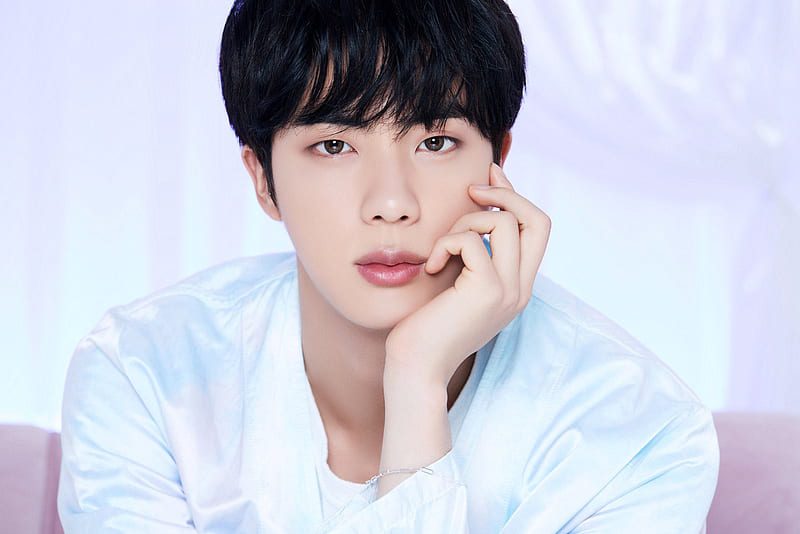A member of a K-pop band wearing a light blue outfit, resting his face on his hand while posing in front of a soft pastel background.