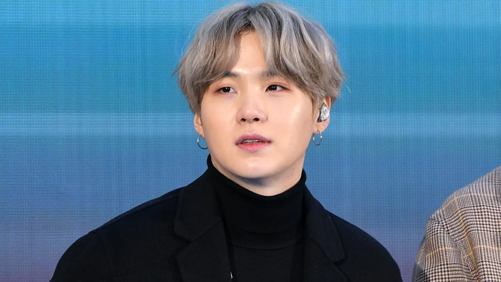 A member of a K-pop band with silver hair, wearing a black turtleneck and coat, looking thoughtful while performing on stage.
