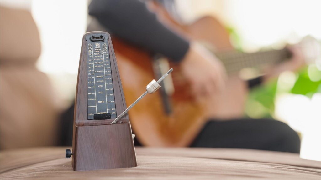 Metronome in focus with guitarist practicing in background, showing importance of timing for improving hand synchronization on guitar