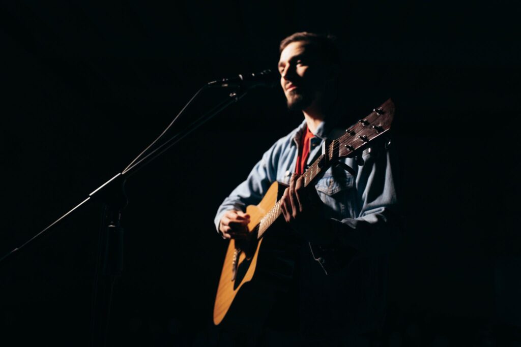 Solo guitarist performing on stage with an acoustic guitar and microphone under dramatic lighting during a live music performance. Listening Skills for Guitarists