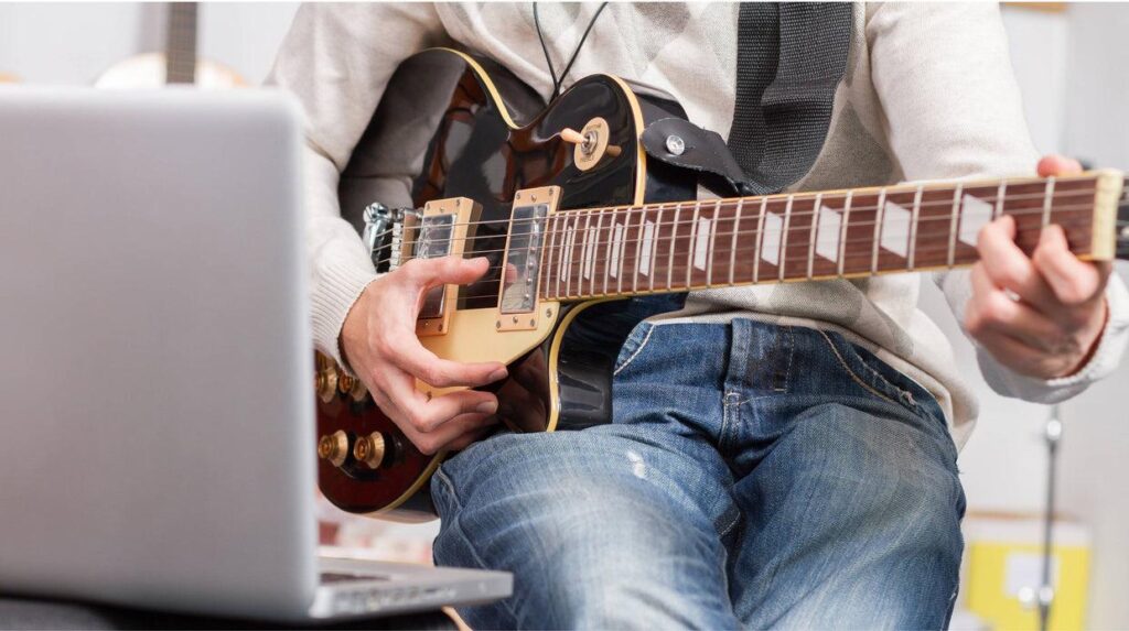 Person practicing electric guitar while using a laptop for online music lessons, guitar learning, or virtual band practice at home.