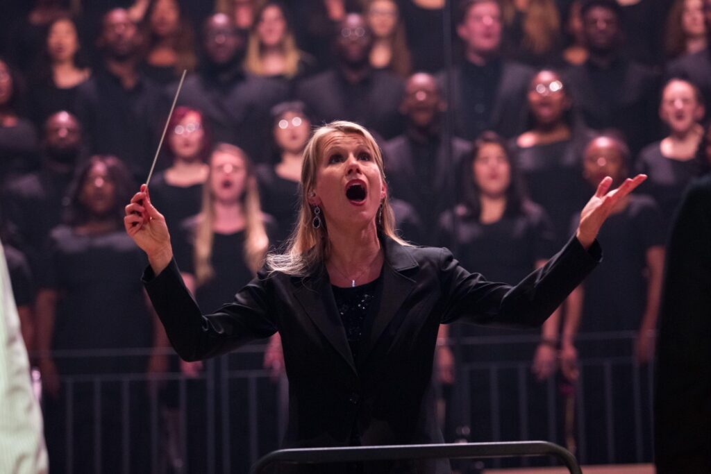 A choir conductor leading a live choral performance with singers in the background, demonstrating the Role of Harmony in Competitive Choir Performances through musical direction, vocal blending, and ensemble coordination.
