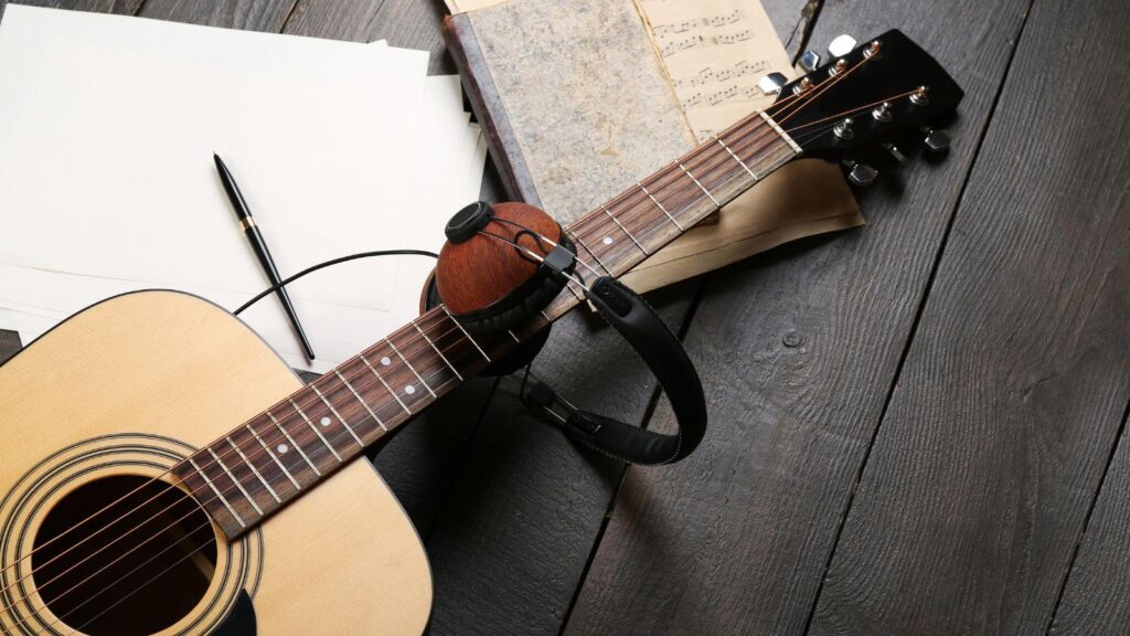 Acoustic guitar with headphones, sheet music, and notebook placed on a wooden table, representing guitar practice, ear training, and music learning. Listening Skills for Guitarists